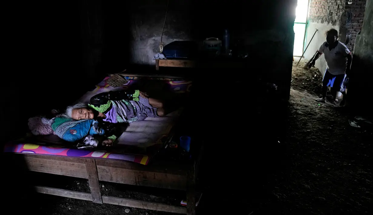 Suratmi yang lumpuh terbaring di tempat tidurnya saat putrinya Ngatiroh (kanan) berjalan di lantai yang telah ditinggikan untuk mencegah air banjir di rumahnya di Timbulsloko, Jawa Tengah, Indonesia, 30 Juli 2022. Ngatiroh mengatakan itu setelah rumah ibunya Suratmi runtuh karena banjir, dia memindahkannya ke rumah berlantai tanah tanpa jendela yang sekarang mereka tinggali. (AP Photo/Dita Alangkara)