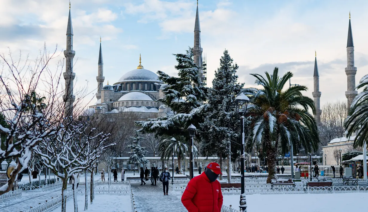 Orang-orang berjalan di alun-alun yang tertutup salju di depan Masjid Biru di Istanbul pada 23 Januari 2022. Badai musim dingin dan hujan salju tetap berlaku di sebagian besar wilayah Turki, menyebabkan penutupan jalan antara ribuan desa dan kota di banyak daerah. (Yasin AKGUL / AFP)