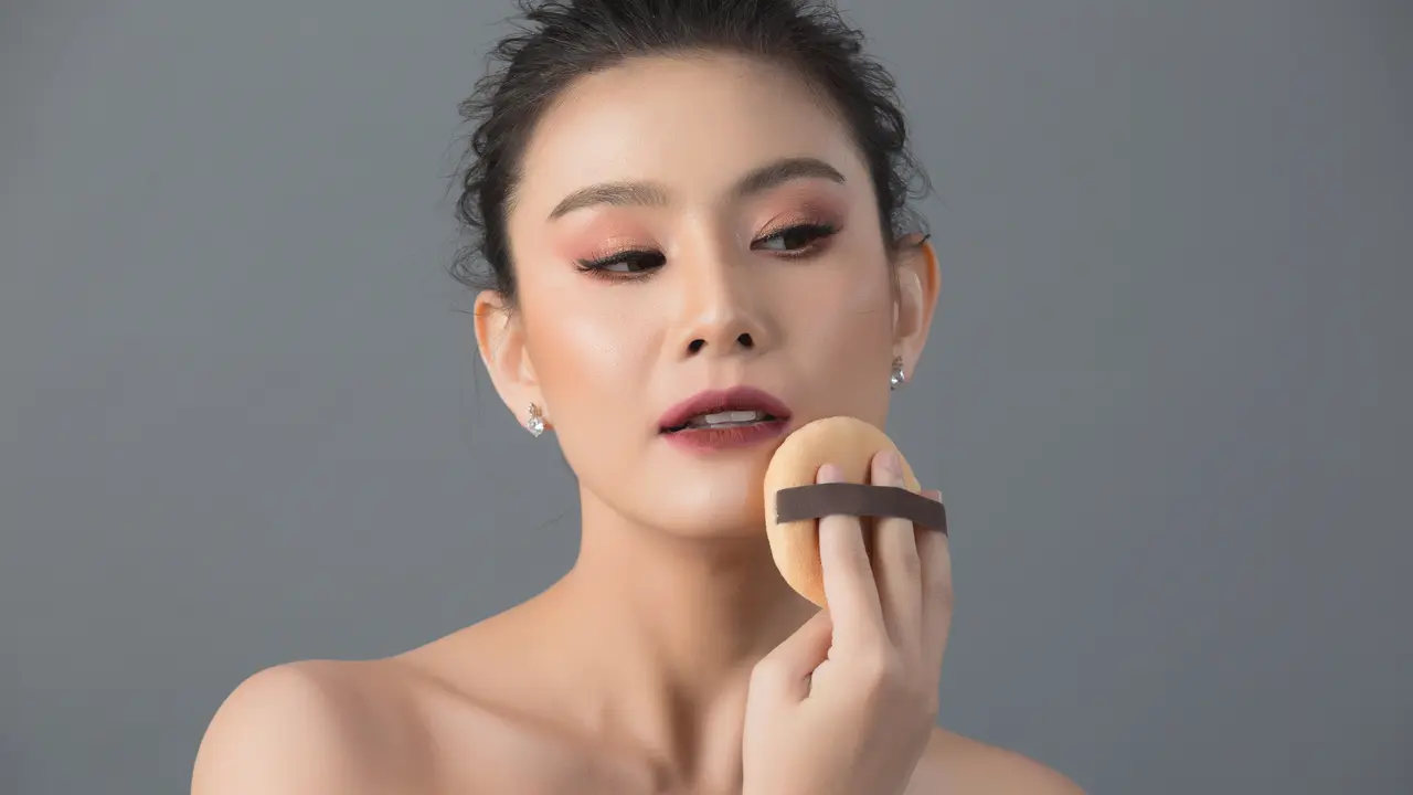 Seorang perempuan sedang mengaplikasikan bedak menggunakan puff ke wajahnya (Foto Dok: Freepik/jcomp)