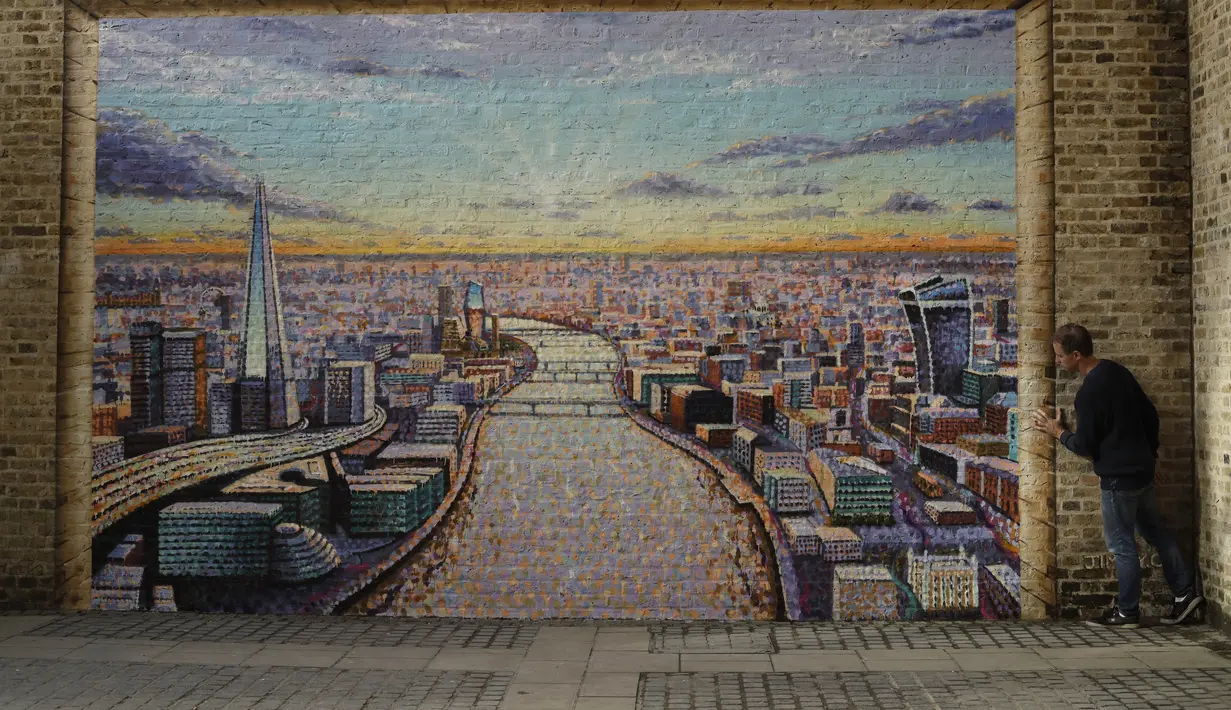 Seniman jalanan Australia James Cochran, berpose untuk mendemonstrasikan efek 3D dari bingkai yang dia lukis agar sesuai dengan tembok bata setelah selesai bekerja membuat mural lanskap kota London di dinding Network Rail oleh Blackfriars Station, di London (5/10/2020). (AP Photo/Matt Dunham)
