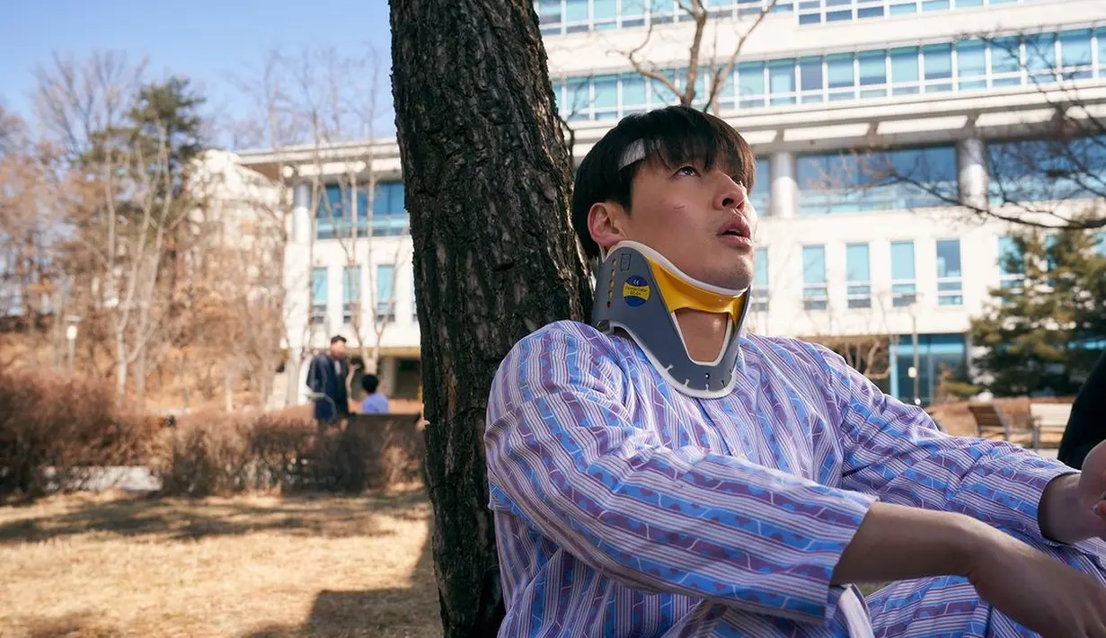 Jung Yeol tampak termenung di bawah pohon dengan baju pasien. Terlihat juga sejumlah luka di tubuhnya. (Foto: Mindmark via Soompi)