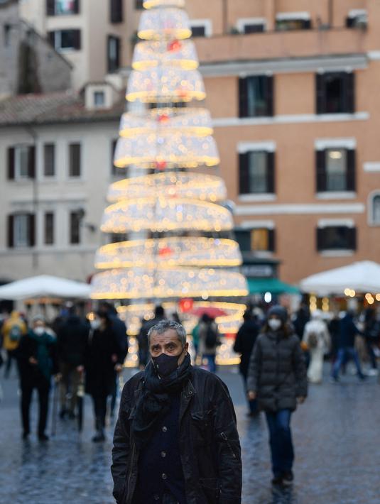Seorang pejalan kaki, mengenakan masker, berdiri di depan pohon natal di jalan Roma, Italia pada Kamis (23/12/2021). Pemerintah Italia telah mewajibkan kembali penggunaan masker di luar ruangan untuk menahan peningkatan kasus Covid-19 yang didorong oleh varian Omicron. (Filippo MONTEFORTE/AFP)