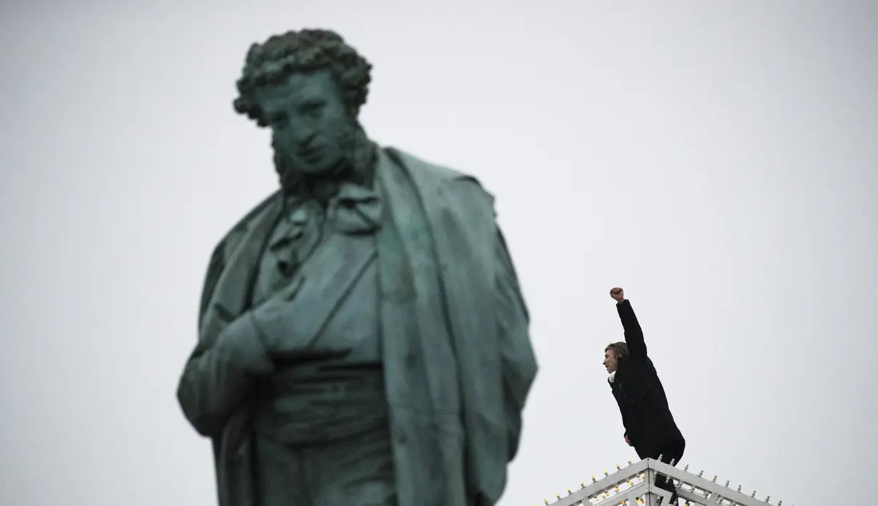 Seorang pria memberi isyarat saat dia memanjat dekorasi Tahun Baru di sebelah patung Alexander Pushkin selama protes terhadap pemenjaraan pemimpin oposisi Alexei Navalny di Moskow, Rusia, Sabtu (23/1/2021). (AP Photo/Pavel Golovkin)