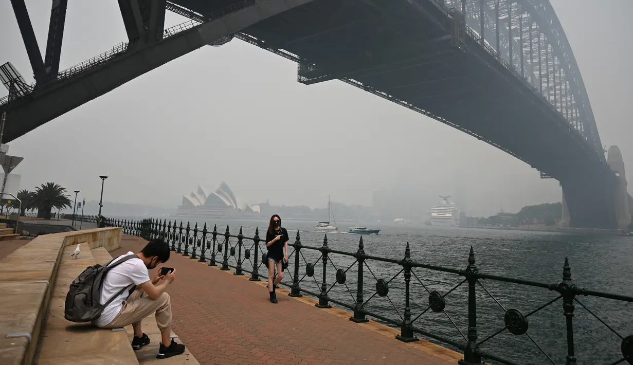 Wisatawan memakai masker saat berpose di bawah Harbour Bridge, Sydney, Australia, Selasa (10/12/2019). Kabut asap yang menyelimuti Sydney dan sebagian wilayah New South Wales dalam beberapa minggu terakhir diperkirakan akan terus berlangsung selama beberapa bulan ke depan. (TAMAN PETER/AFP)