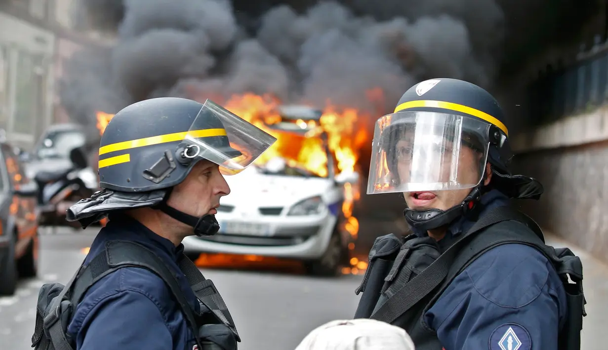 Dua petugas kepolisian berada di dekat mobil dinas yang terbakar akibat demo buru di Paris, Prancis (18/5). Demo buruh tersebut menentang kekerasan polisi dan melawan reformasi hukum yang berakhir bentrok dengan polisi. (REUTERS / Charles Platiau)