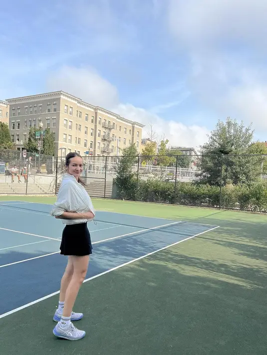 Gaya sporty Enzy Storia di lapangan tenis. Ia mengenakan cropped jacket tenis berwarna putih, dipadu rok hitam, dan sneakers putih. [Foto: Instagram/enzystoria]