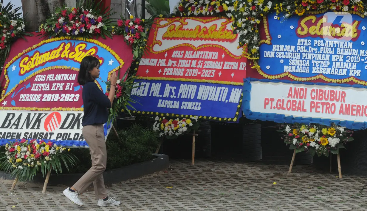 Seorang perempuan bermain ponsel di dekat  karangan bunga ucapan selamat yang berjejer di sekitar Gedung Merah Putih KPK, Jakarta, Jumat (20/12/2019). Papan karangan bunga tersebut berisi ucapan selamat terhadap dilantiknya pimpinan KPK periode 2019-2023. (merdeka.com/Imam Buhori)