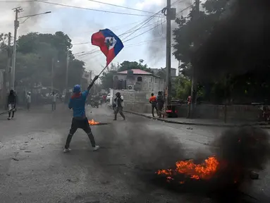 Seorang pria memegang bendera Haiti sementara ban terbakar saat demonstrasi menentang ketidakamanan di Carrefour-Feuilles, Distrik Port-au-Prince, Haiti, 14 Agustus 2023. (Richard PIERRIN/AFP)