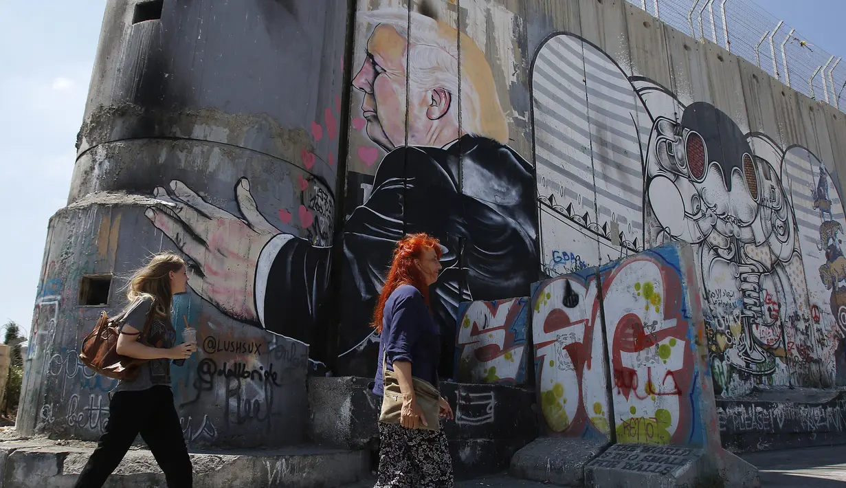 Wisatawan berjalan melewati grafiti yang menggambarkan Presiden AS Donald Trump di tembok pemisah Israel - Palestina di kota Bethlehem (4/8). Tembok ini memisahkan antara Israel dan Palestina. (AFP Photo/Musa Al Shaer)