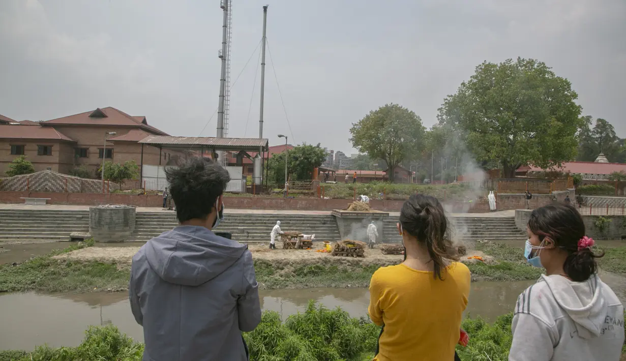Anggota keluarga mengawasi di seberang sungai Bagmati ketika pria Nepal dengan pakaian pelindung mengkremasi mayat korban COVID-19 di dekat kuil Pashupatinath di Kathmandu, Nepal (3/5/2021). (AP Photo/Niranjan Shrestha)