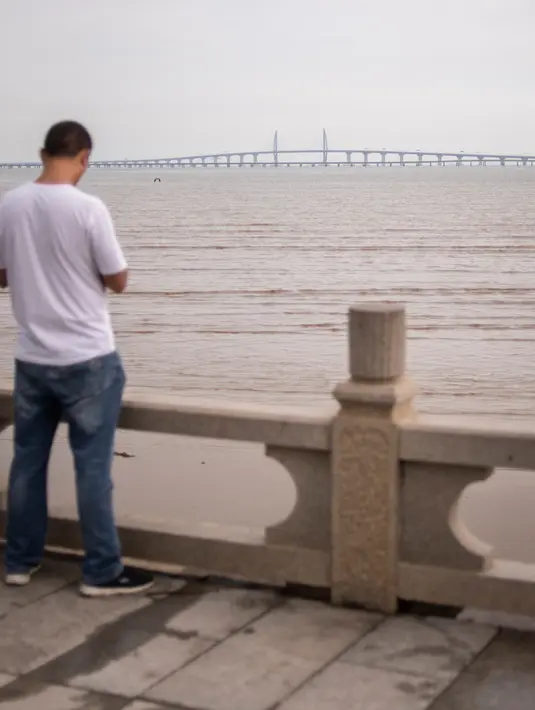 Orang-orang berdiri di pinggir pantai menghadap Jembatan Hong Kong-Zhuhai-Makau (HKZM) di Zhuhai (22/10). Jembatan laut terpanjang di dunia yang menghubungkan Hong Kong, Makau, dan daratan Cina ini akan dibuka 24 Oktober 2018. (AFP Photo/Fred Dufour)