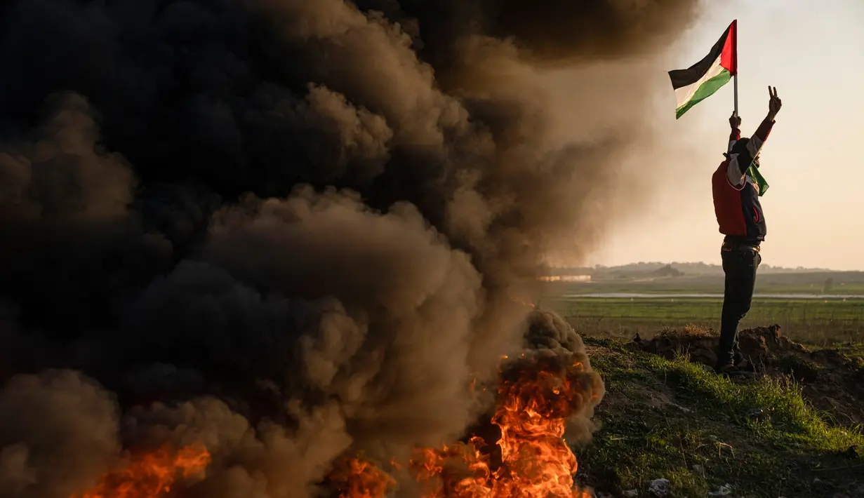 Warga Palestina membakar ban dan mengibarkan bendera nasional saat protes terhadap serangan militer Israel terhadap kamp pengungsi Jenin di perbatasan Israel-Palestina, sebelah timur Kota Gaza, 26 Januari 2023. Presiden Palestina Mahmoud Abbas mengumumkan masa berkabung tiga hari, di mana bendera akan dikibarkan setengah tiang. (AP Photo/Fatima Shbair)