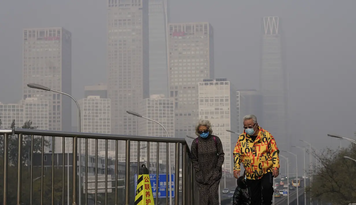 Sepasang suami istri yang mengenakan masker untuk membantu mengekang penyebaran virus corona berjalan di jembatan di atas gedung perkantoran di Kawasan Pusat Bisnis yang diselimuti kabut polusi di Beijing, Kamis (18/11/2021).  (AP Photo/Andy Wong)