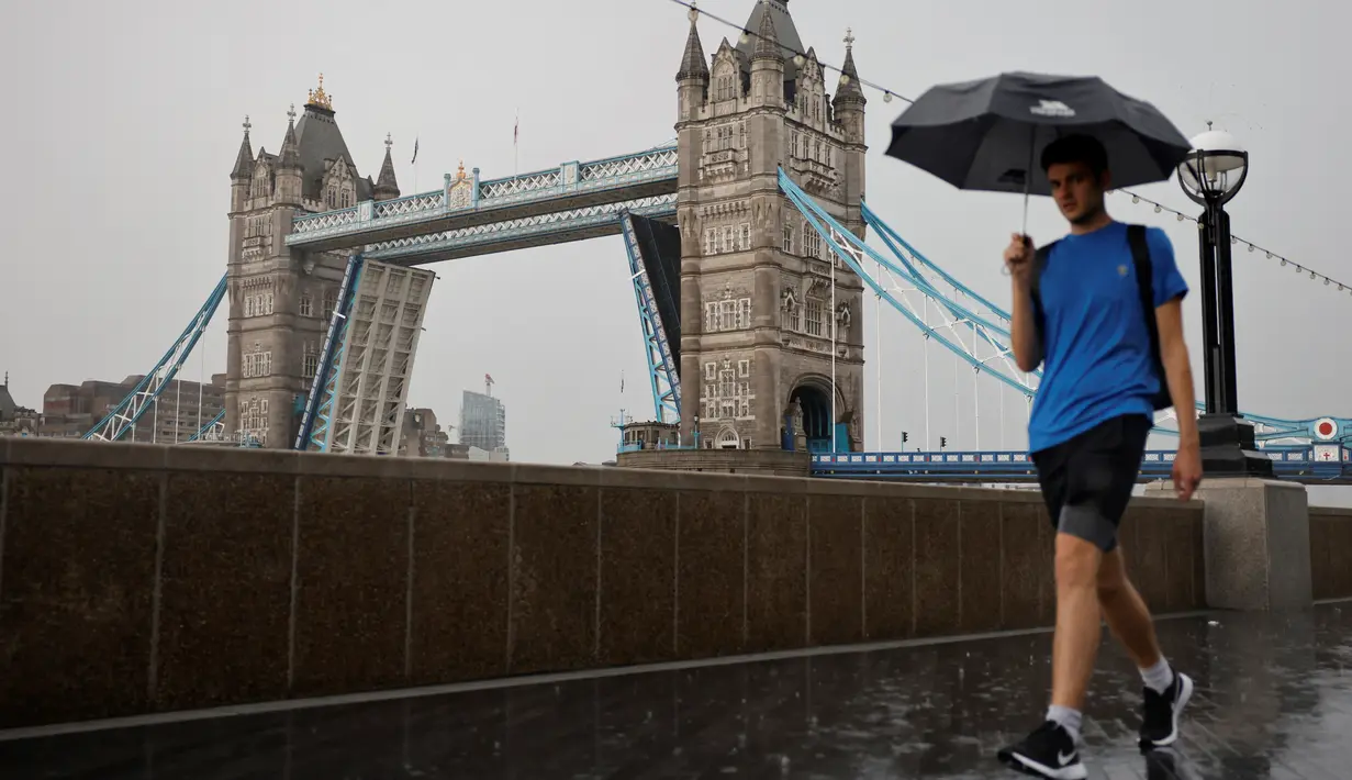 Pejalan kaki berjalan di tengah hujan di tepi sungai Thames saat Tower Bridge ondonterjebak dalam posisi terbuka, Senin (9/8/2021). Tower Bridge London macet terbuka mengakibatkan gangguan lalu lintas di kedua sisi Sungai Thames karena mobil dan pejalan kaki tidak dapat menyeberang (Tolga Akmen/AFP)