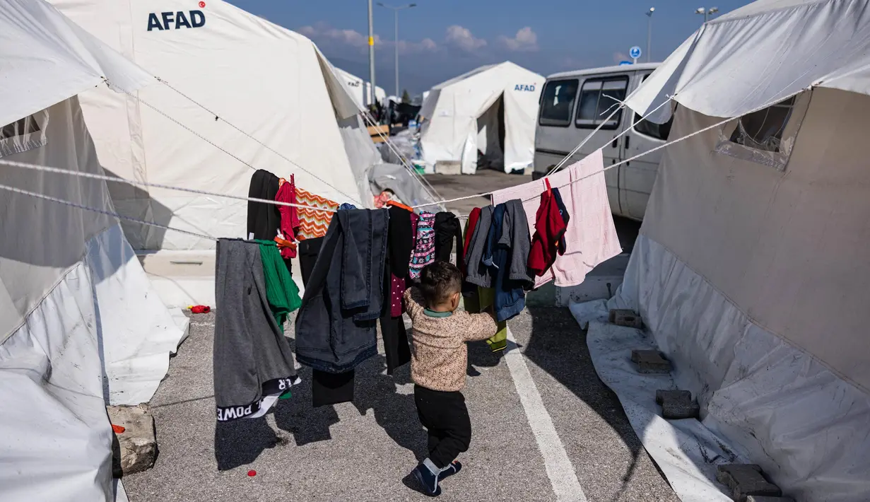 Seorang anak laki-laki berjalan di dekat pakaian yang digantung untuk dijemur di sebuah kamp darurat di sebelah stadion Hatay yang baru di Antakya, Turki Selatan, pada 22 Februari 2023. Pemerintah Turki menjadikan kompleks stadion sebagai tempat pengungsian untuk menampung ribuan korban gempa yang kehilangan rumahnya. (Sameer Al-DOUMY/AFP)