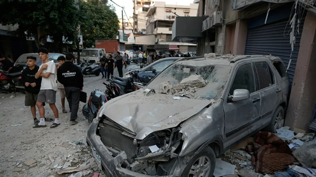 Orang-orang melintas di dekat sebuah mobil yang rusak di lokasi serangan Israel yang menghantam sebuah gedung apartemen di Dahiyeh, wilayah pinggiran selatan Beirut, Lebanon, Minggu, (23/11/2025).