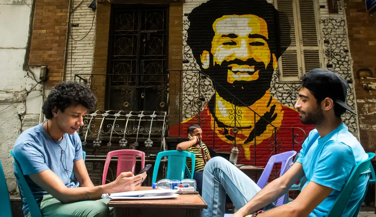 Dua pemuda duduk bersantai dengan latar belakang mural bintang Liverpool, Mohamed Salah di sebuah kedai kopi di pusat kota Kairo, 30 April 2018. Mural pemain timnas Mesir tersebut menghiasi sudut negara asalnya. (AFP Photo/KHALED DESOUKI)