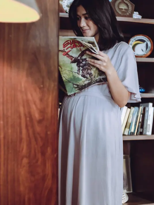 Ini salah satu adegan dalam film Inang. Memerankan Wulan dengan usia kandungan menua, Naysilla Mirdad tampil dengan terusan longgar lengan pendek beraksen pita di bagian perut. Rambutnya tergerai sementara perut tampak membuncit. Wulan dengan air muka tersenyum tampak asyik membaca buku dengan latar rak berisi sejumlah bacaan dan aksesoris. (Foto: Dok. IDN Pictures)
