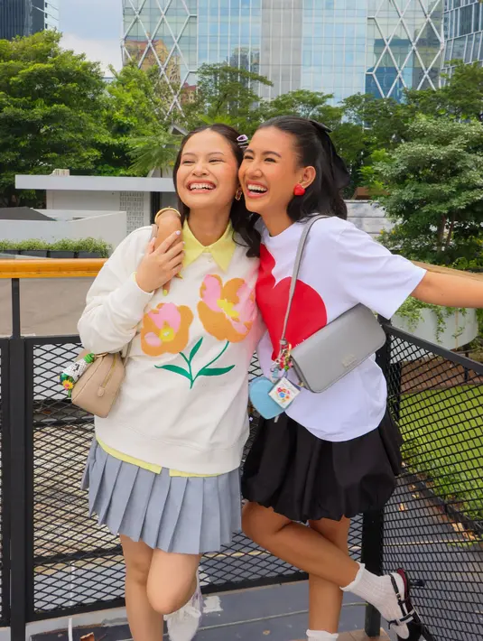 Pada momen lainnya, mereka tampak menikmati suasana sore hari dari atas rooftop, tetap dengan tampilan yang stylish. Kali ini, Hanggini memilih tampilan preppy style, dengan perpaduan white sweater dan mini pleated skirt. Ia pun mengenakan tas yang sama seperti Shenina di tampilan sebelumnya namun dalam warna berbeda, yaitu Grace East-West Shoulder Bag berwarna taupe.  [Kate Spade]