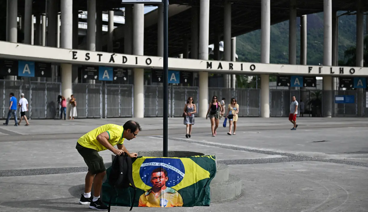 <p>Seorang pria memasang bendera Brasil dengan gambar mendiang legenda sepak bola Pele di bawah tanda jalan bertuliskan "Avenue King Pele" di luar stadion Maracana di Rio de Janeiro, Brasil pada 4 Januari 2023. Balai Kota Rio de Janeiro secara resmi menamai jalan yang mengelilingi stadion Maracana Avenue Rei Pele (King Pele). (MAURO PIMENTEL/AFP)</p>