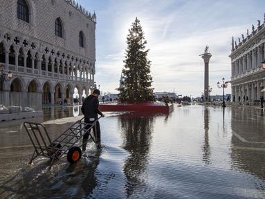 Seorang porter mengarungi St Mark's Square  yang dibanjiri air laut di samping pohon Natal di Venesia, Italia, Sabtu (4/12/2021). Air mencapai 99 sentimeter di atas permukaan laut dan bagian terendah kota tenggelam. (AP Photo/Luigi Costantini)
