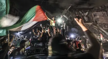 Warga mengibarkan bendera Palestina saat merayakan gencatan senjata antara Israel dengan Hamas di depan bangunan yang hancur di Kota Gaza, Palestina, Jumat (21/5/2021). Israel melakukan gencatan senjata dengan gerakan Islam yang berkuasa di Jalur Gaza, Hamas. (MAHMUD HAMS/AFP)