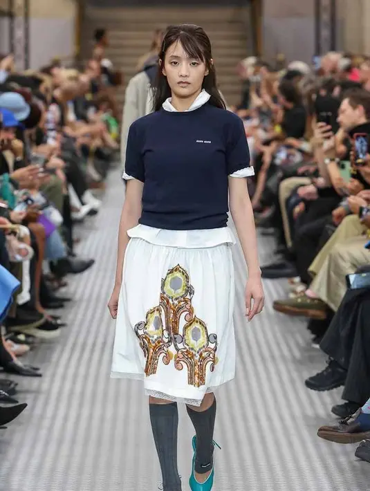 Pertama ada sang brand ambassador Minnie (G)I-DLE. Idol 26 tahun mencuri perhatian dengan gaya femininnya berupa rok putih, kaus Miu Miu yang berkelas dan kemeja putih. Penampilan perdana Minnie di panggung peragaan busana dilengkapi dengan kaus kaki setinggi lutut dan sepatu hak tinggi berwarna biru kehijauan