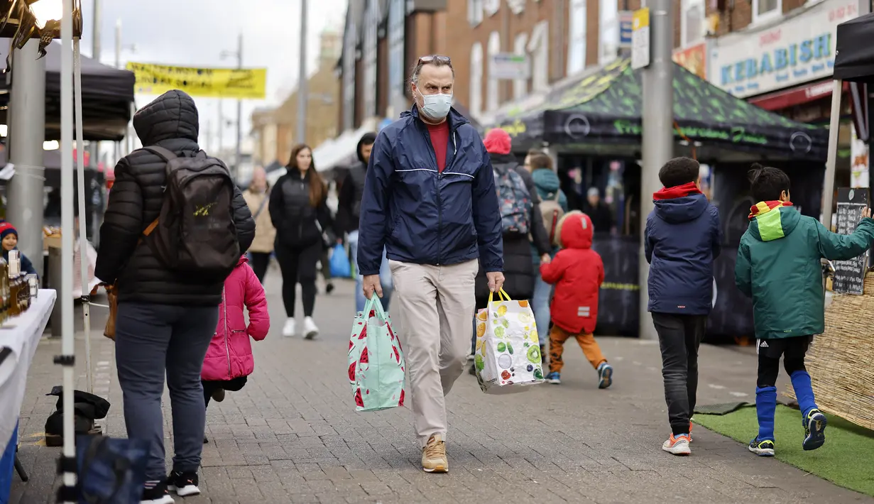 Seorang pria yang mengenakan masker membawa tas belanjaan saat melewati kios-kios di Walthamstow Market, London, Minggu (22/11/2021). Indeks Harga Konsumen Inggris naik ke angka 4,2 persen dalam 12 bulan hingga Oktober 2021, mencapai level tertinggi sejak November 2011. (Tolga Akmen/AFP)