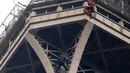 Petugas pemadam kebakaran berusaha mendekati seorang pria (baju hitam di bawah) yang memanjat Menara Eiffel tanpa peralatan keselamatan di Paris, Senin (20/5/2019). Akibat ulahnya, Menara Eiffel ditutup dan pihak pengelola melakukan evakuasi dan pengamanan. (AP/Michel Euler)