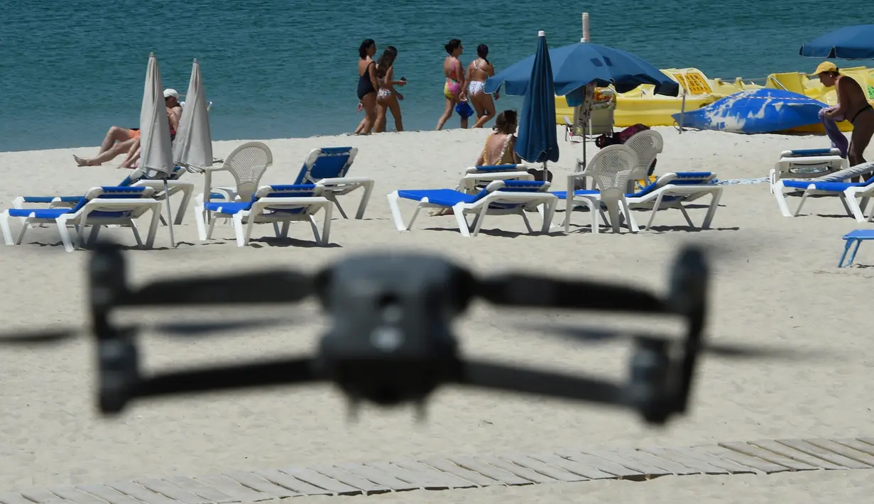 Sebuah drone atau pesawat tak berawak yang diterbangkan oleh polisi setempat memperingatkan orang-orang untuk menjaga jarak di pantai Areas di Sanxenxo, Spanyol, Selasa (14/7/2020). Drone tersebut untuk memantau jaga jarak antarwisatawan saat berlibur di masa pandemi Corona. (MIGUEL RIOPA / AFP)