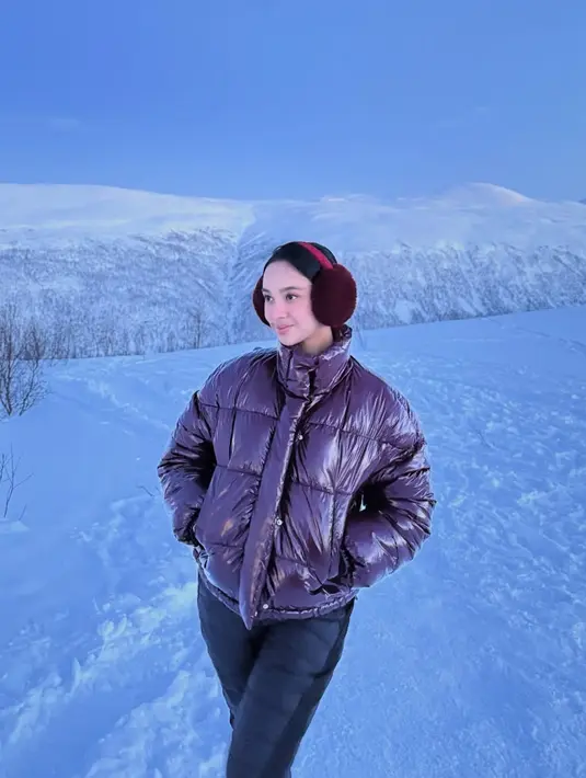 Lyodra mengenakan penutup telinga maroon dipadukan jaket padding model pendeknya. Dipadukan bawahan celana hitam. [@lyodraofficial]