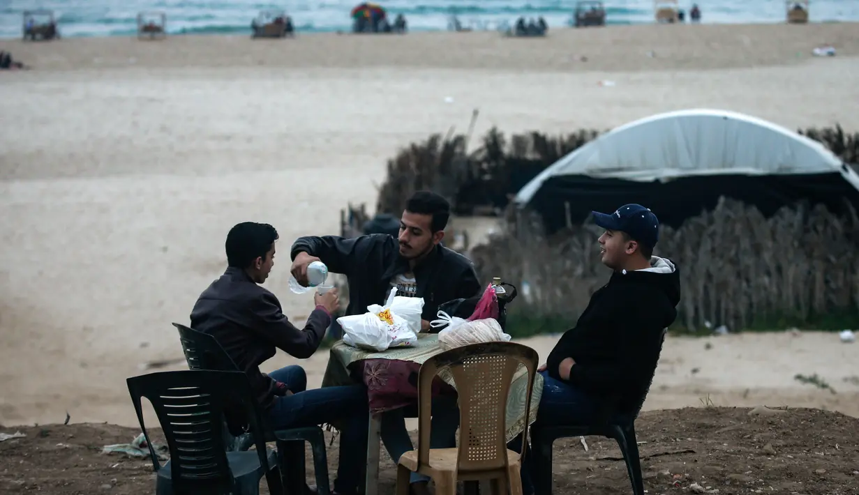 Warga Palestina berkumpul untuk berbagi makanan buka puasa selama bulan suci Ramadan, saat pandemi virus corona, di sepanjang pantai kota Gaza pada 13 Mei 2020. Mereka menunggu waktu berbuka puasa sambil menyaksikan matahari terbenam. (Photo by MAHMUD HAMS / AFP)