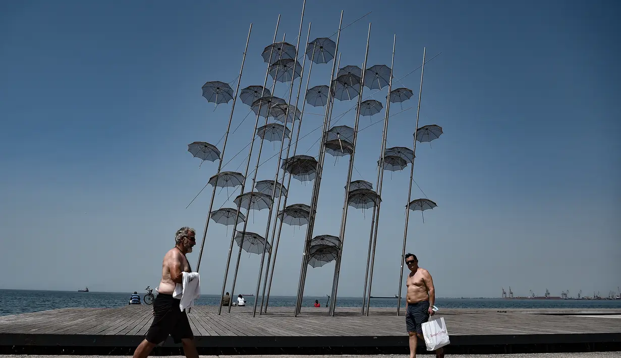Orang-orang berjalan melewati instalasi ikonik "Umbrellas" karya pematung, pelukis, dan arsitek Yunani George Zongolopoulos, di tepi pantai Thessaloniki (23/6/2021).  Yunani bersiap menghadapi gelombang panas pertama musim panas. (AFP/ Sakis Mitrolidis)