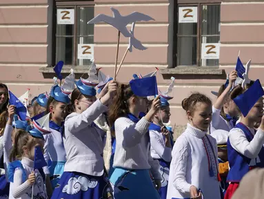 Anak-anak berbaris dengan latar belakang lembaran yang yang dipajang di jendela menggambarkan huruf Z simbol militer Rusia selama perayaan memperingati 318 tahun kota Kronstadt di luar St. Petersburg, Rusia (21/5/2022). (AP Photo/Dmitri Lovetsky)