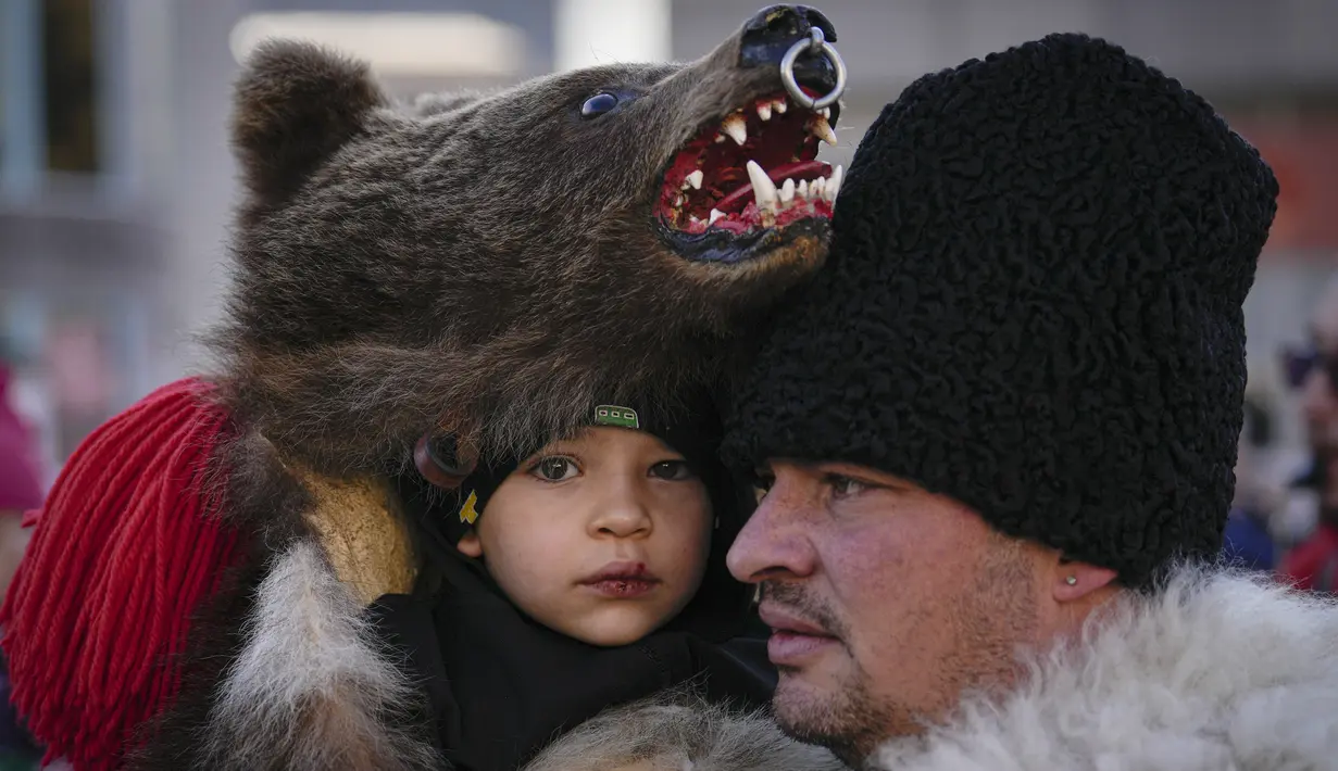 Seorang pria menggendong anak kecil yang mengenakan kostum bulu beruang saat parade di Comanesti, Rumania, Jumat, 30 Desember 2022. Penduduk setempat khawatir tradisi itu akan hilang karena sejumlah besar pemuda Romania telah meninggalkan daerah itu untuk mencari kehidupan yang lebih baik di negara-negara Uni Eropa yang lebih kaya. (AP Photo/Vadim Ghirda)