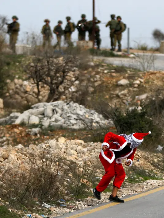 Seorang demonstran Palestina mengenakan kostum Santa Claus melempar batu ke arah pasukan keamanan Israel saat terjadi bentrokan di pos pemeriksaan Atarot di pinggiran utara Yerusalem (19/12). (AFP Photo/Abbas Momani)