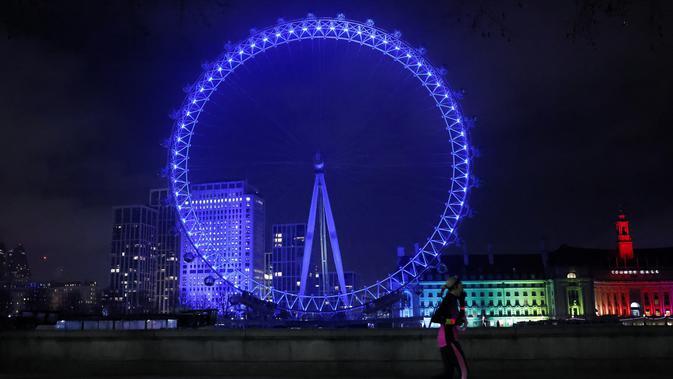 Pejalan kaki melintas di depan London Eye yang diterangi dengan cahaay biru untuk berterima kasih kepada National Health Service (NHS) di London, Kamis (7/1/2021). NHS merupakan sistem layanan kesehatan nasional yang menjadi ujung tombak Inggris dalam memerangi pandemi Covid-19. (Tolga Akmen / AFP)