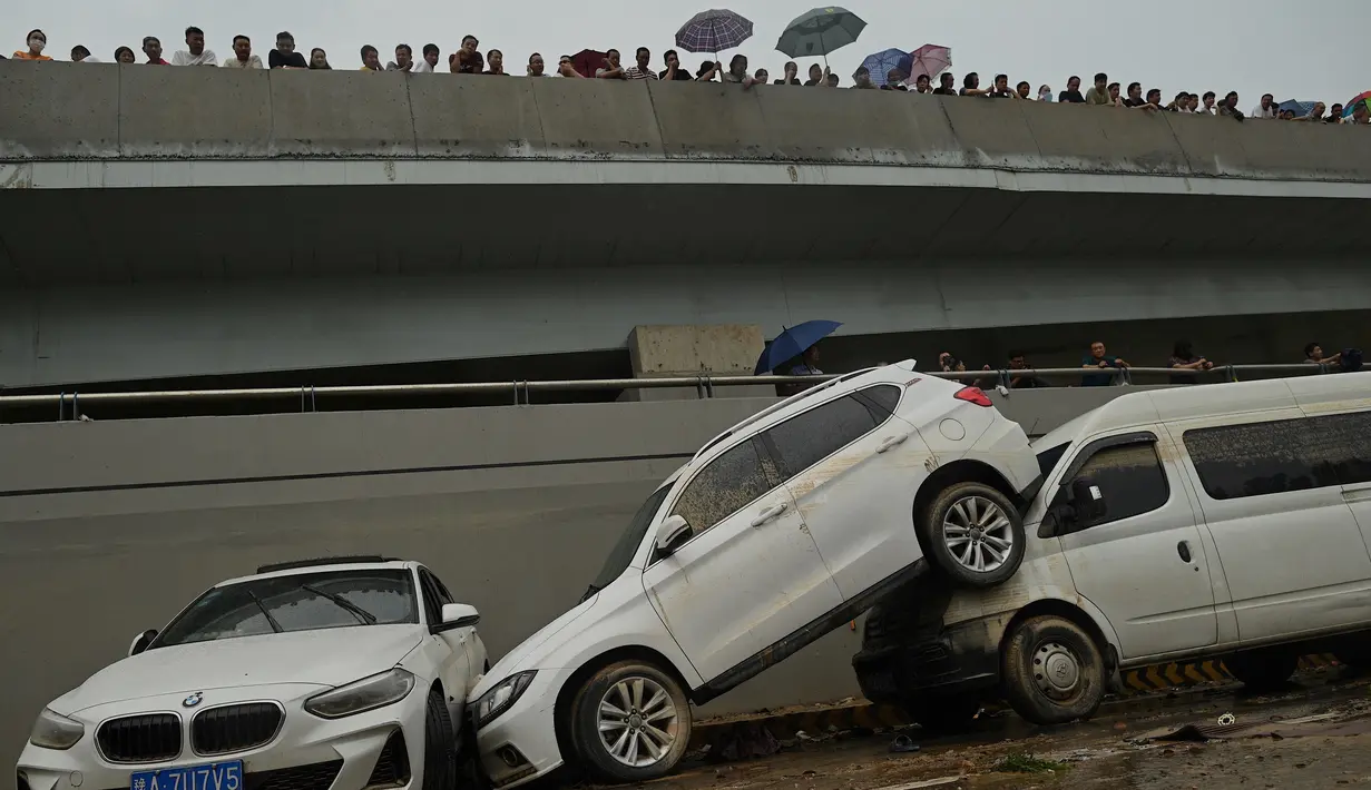 Orang-orang melihat mobil-mobil yang terendam banjir setelah hujan lebat melanda Zhengzhou di provinsi Henan, China, Kamis (22/7/2021). Curah hujan yang tinggi mengakibatkan aliran air sungai meluap ke jalan dan membuat sejumlah kendaraan terbawa arus dan bertumpuk di sudut jalan. (Noel Celis/AFP)