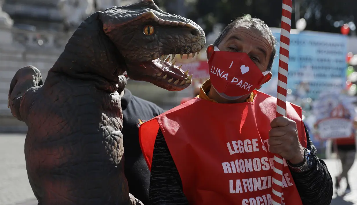 Pekerja taman memakai masker untuk mencegah COVID-19 saat ia berpura-pura digigit dinosaurus saat menghadiri demonstrasi pekerja sirkus yang menuntut dukungan pemerintah Italia karena aktivitas mereka ditutup sejak awal wabah COVID-19, di Roma (26/3/2021). (AP Photo/Gregorio Borgia)