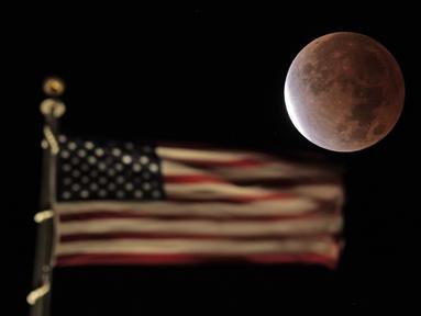 Bayangan bumi menutupi bulan purnama selama gerhana bulan sebagian atau parsial saat terbenam melewati bendera AS di atas sebuah gedung di pusat kota Kansas City, Missouri, Jumat (19/11/2021). (AP Photo/Charlie Riedel)