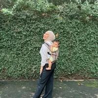 Di foto terbaru yang diunggahnya, Irish Bella tampak menggendong anak keduanya di punggungnya. Ia mengenakan atasan berwarna putih, dipadu dengan celana panjang hitam bergaris, dan hijab cokelat muda. Sang buah hati tampak gemas dengan outfit berwarna kuning. Foto: Instagram.