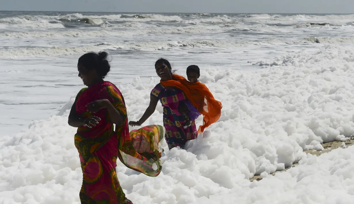 Penduduk setempat berjalan di atas limbah busa yang disebabkan polutan saat bercampur dengan ombak di pantai di Chennai (29/11/2019). Limbah busa tersebut  membuat penduduk dan pengunjung pantai merasa sakit. (AFP/Arun Sankar)
