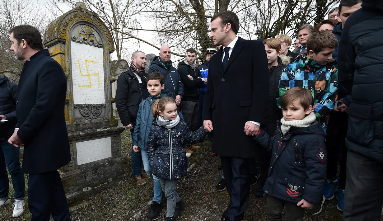 Presiden Prancis Emmanuel Macron berjalan melewati makam Yahudi yang dicoret simbol Nazi di pemakaman Yahudi, Quatzenheim, Prancis, Selasa (19/2). Macron menyempatkan diri untuk memeriksa kerusakan yang ditimbulkan. (Frederick FLORIN/AFP)