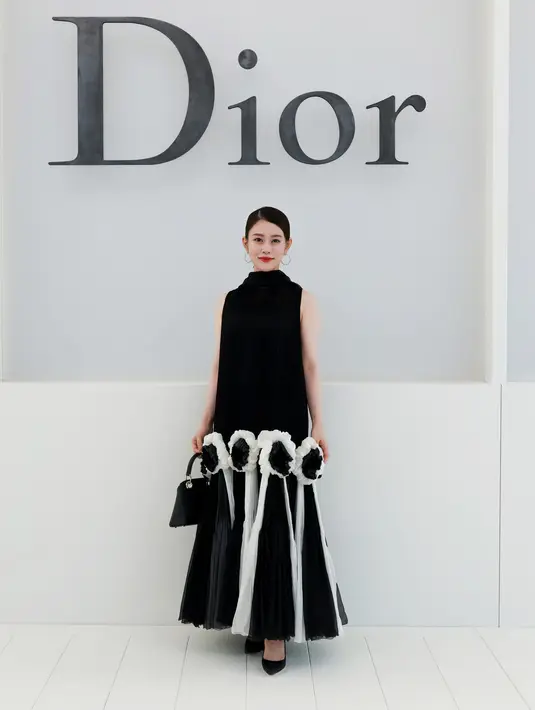 Mitsuki Takahata memilih dress pleated hitam dengan detail bunga yang statement. Dipadukan anting hoop dan tas mini hitam [dok. Dior]
