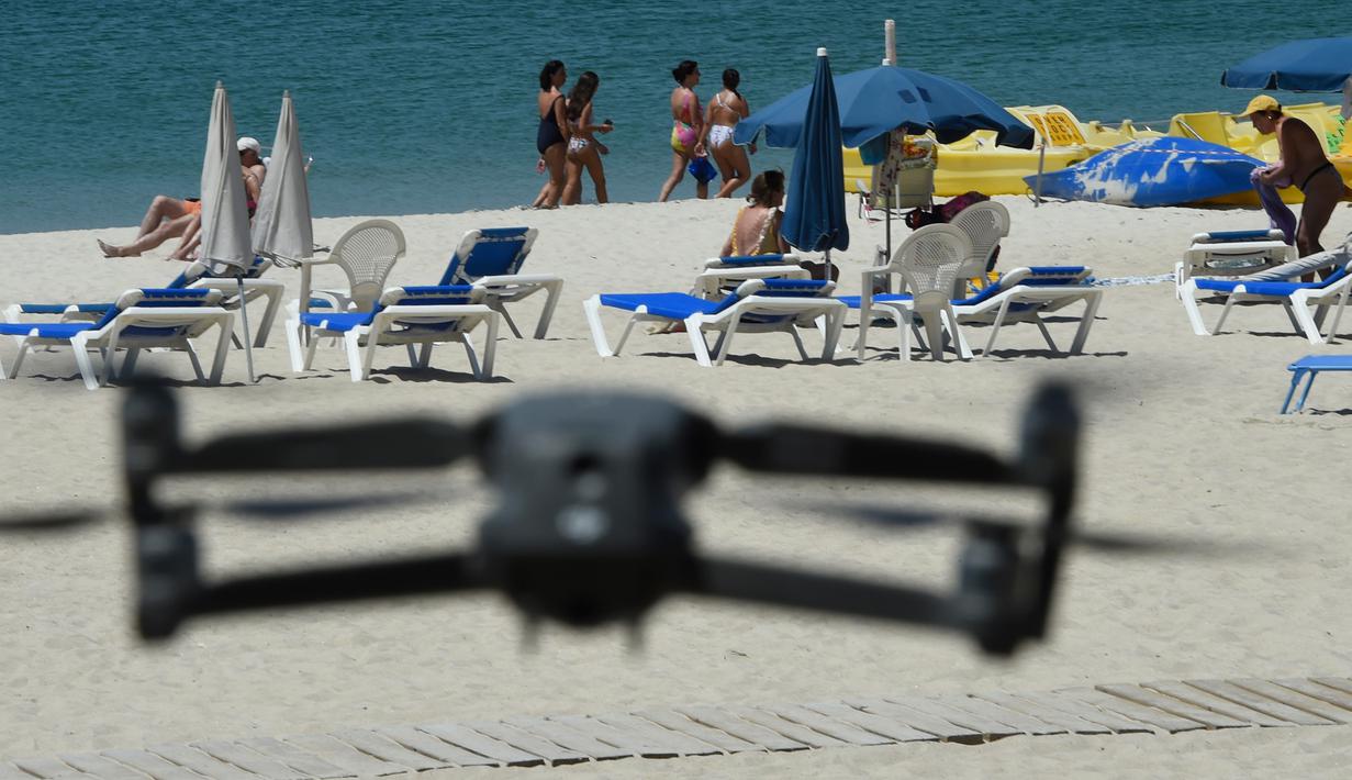 Sebuah drone atau pesawat tak berawak yang diterbangkan oleh polisi setempat memperingatkan orang-orang untuk menjaga jarak di pantai Areas di Sanxenxo, Spanyol, Selasa (14/7/2020). Drone tersebut untuk memantau jaga jarak antarwisatawan saat berlibur di masa pandemi Corona. (MIGUEL RIOPA / AFP)
