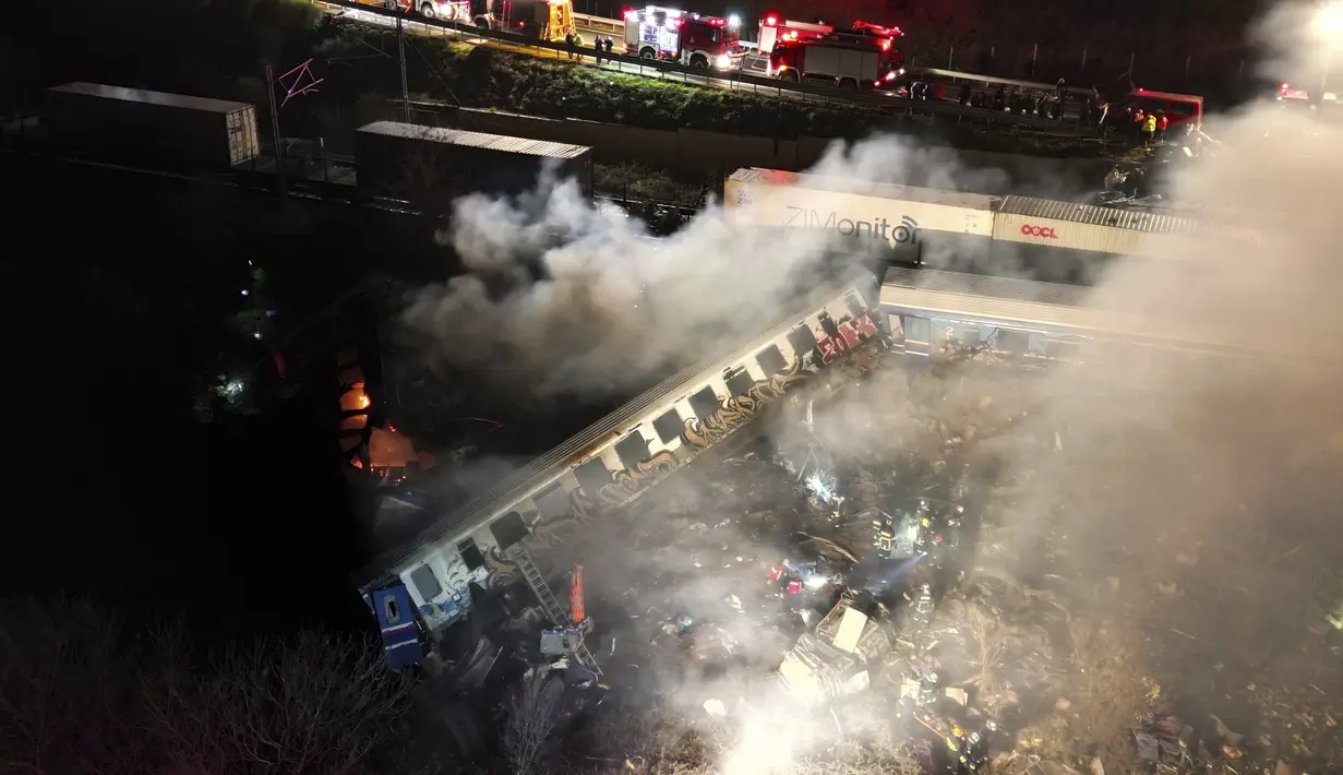 Asap mengepul dari kereta saat petugas pemadam kebakaran dan penyelamat beroperasi setelah tabrakan di dekat kota Larissa, Yunani, Rabu dini hari (1/3/2023). Petugas penyelamat terlihat membawa alat penerang di gerbong-gerbong mencari penumpang yang terjebak. (AP Photo/Vaggelis Kousioras)