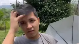 Penampilannya begitu sederhana dengan kaos abu-abu longgar dan celana hitam santai. Sambil berjalan di antara pepohonan karet, Jirayut menunjukkan bagaimana proses penyadapan getah dilakukan. Sesekali, ia bercanda dengan menunjukkan wadah kecil berisi getah karet, membuat suasana terasa akrab dan ceria. (Liputan6.com/IG/@jirayutdaa4official)