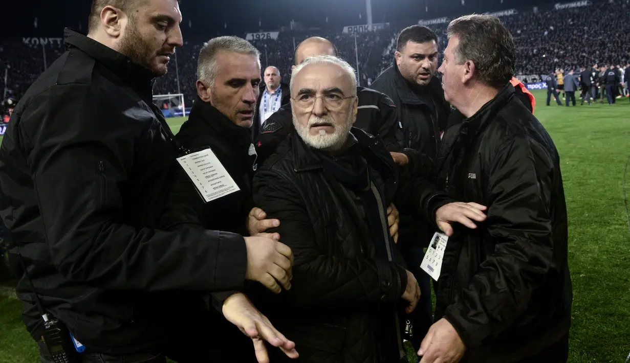 Presiden PAOK Ivan Savvidis diamankan petugas setelah turun ke lapangan dengan pistol di pinggangnya saat Liga Super Yunani di Stadion Toumbas, Minggu (11/3). Laga yang mempertemukan PAOK dengan AEK Athens terpaksa harus ditunda. (SAKIS MITROLIDIS/AFP)