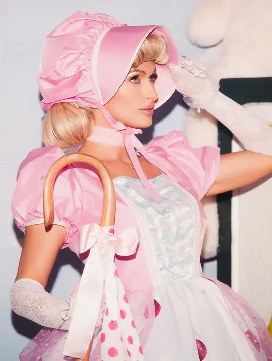 Wajah Paris Hilton dipulas dengan soft glam bernuansa pinkish. Sementara rambut pirangnya ditekuk di balik penutup kepala Bo Peep dan poni yang digulung [@parishilton]