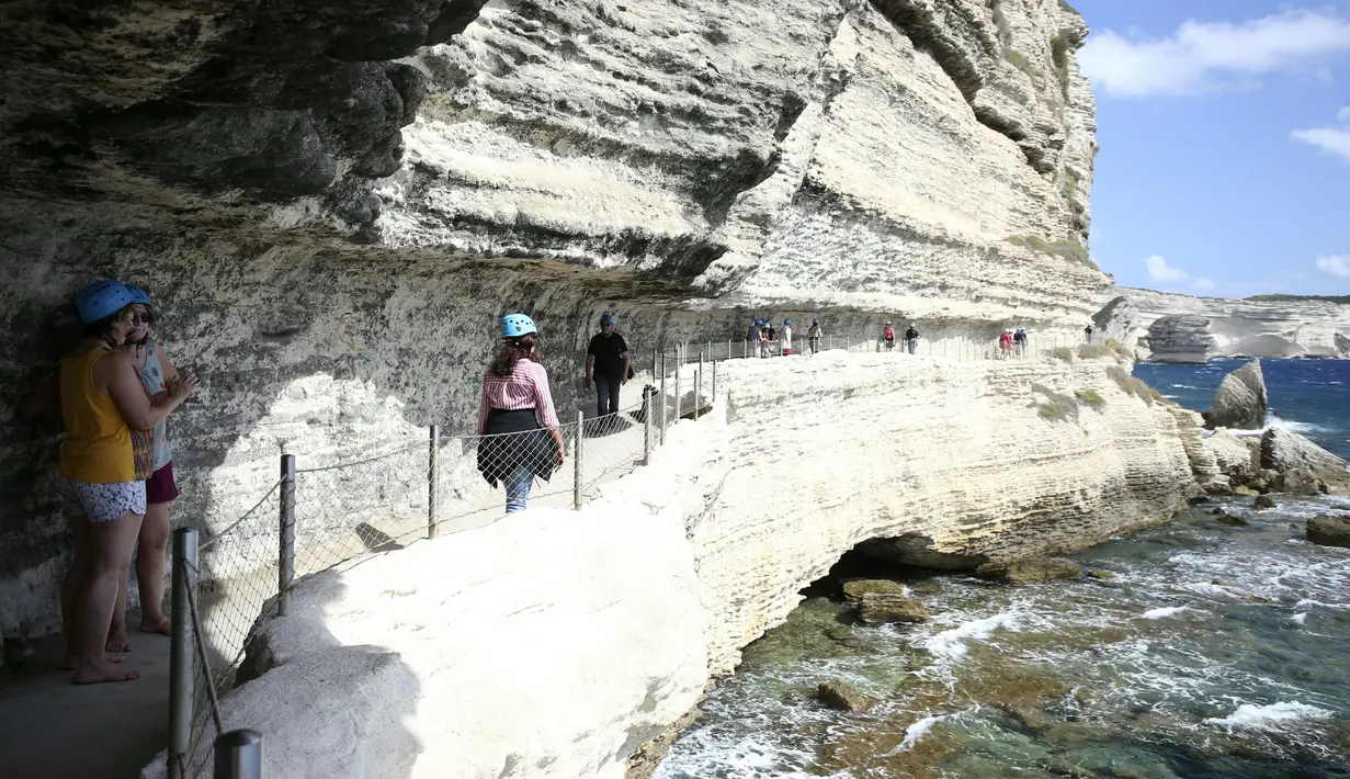 Turis menyusuri jalan setapak di kaki tebing sebelum menaiki tangga Raja Aragon di Bonifacio di pulau Corsica, Mediterania Prancis (19/9/2021). Tangga Raja Aragon digali dengan tangan di tebing batu kapur di suatu malam oleh pasukan Raja Aragon tahun 1420. (AFP/Pascal Pochard-Casabianca)